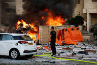 A man stands facing a burning building next to a white car.